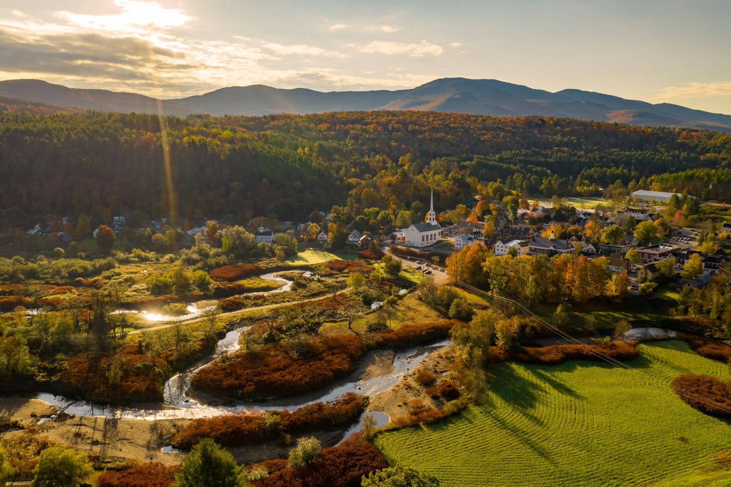 A small New England town with a white church steeple, a river, and autumn foliage below wooded mountains at golden hour